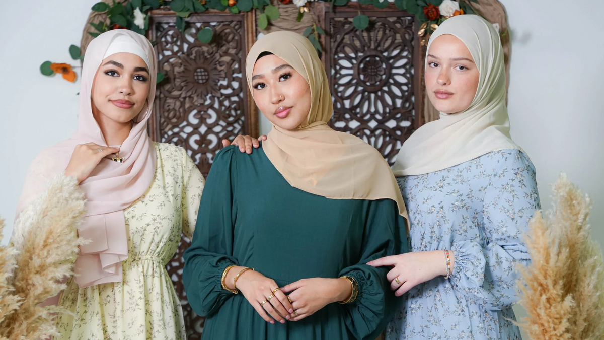 Three women wearing elegant dresses with neutral hijabs against a decorative background, showcasing diverse fashion styles.