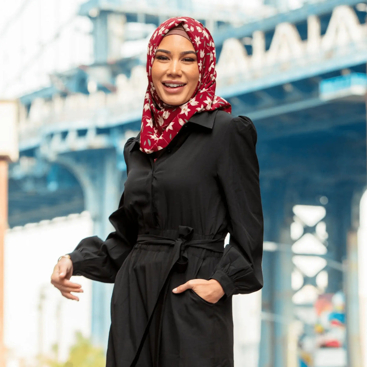 Woman wearing a black modest jumpsuit with a red hijab, standing in an urban setting. Modest Jumpsuits collection by Urban Modesty.