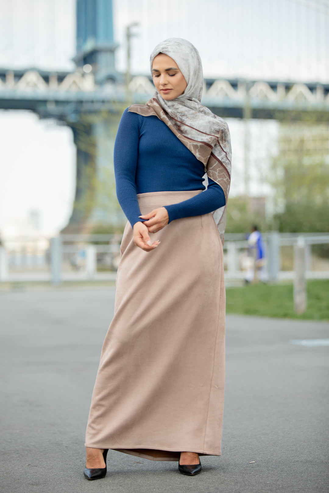 Woman wearing Sand Suede Pencil Maxi Skirt with a blue top and scarf, posing outdoors with a bridge in the background.