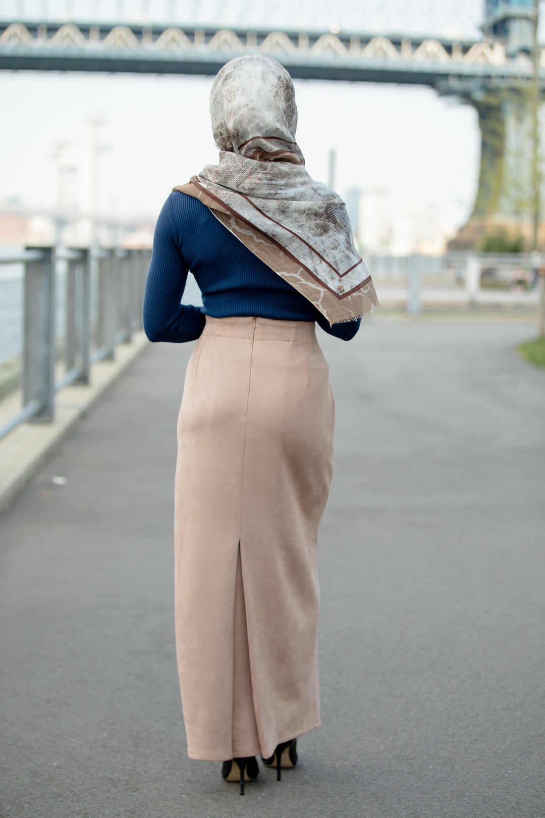 Woman wearing sand suede pencil maxi skirt with back pleat, paired with a blue top and scarf, standing on a pathway near a bridge.