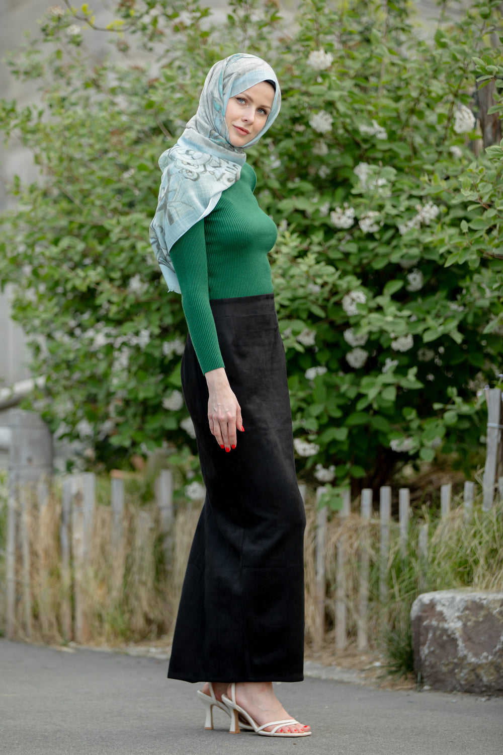 Woman wearing black suede pencil maxi skirt with green top and hijab, standing outdoors in a garden