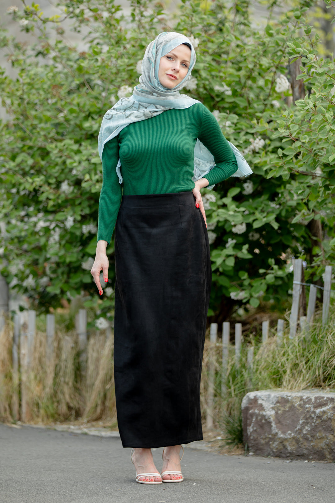 Woman wearing Black Suede Pencil Maxi Skirt, green top, and hijab standing in front of greenery.