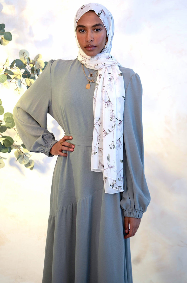 Woman wearing gray asymmetrical hem maxi dress with cuff sleeves and white chiffon hijab, featuring ruffles at hem and sleeves.