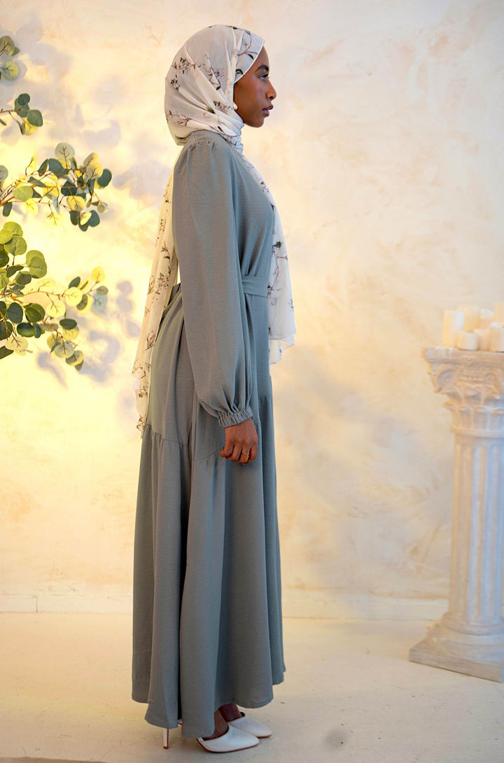 Woman wearing gray asymmetrical hem maxi dress with cuff sleeves, paired with a floral chiffon hijab and white heels, side profile view.