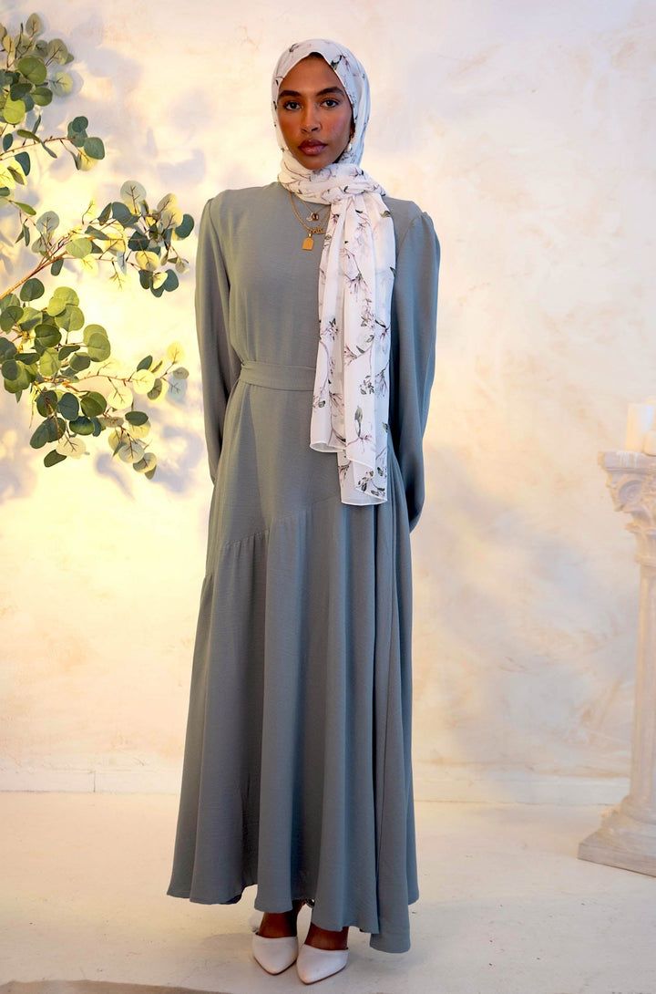 Woman wearing Gray Asymmetrical Hem Maxi Dress with cuff sleeves, paired with Chiffon hijab and white shoes, in well-lit indoor setting.