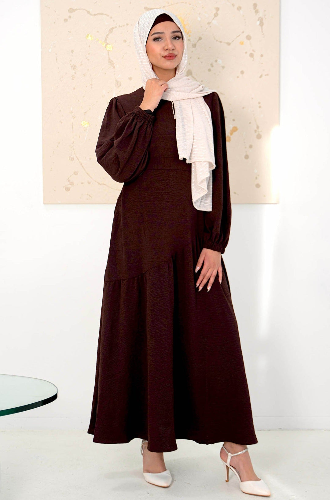 Woman wearing brown asymmetrical hem maxi dress with cuff sleeves, paired with a white chiffon hijab and white heels.