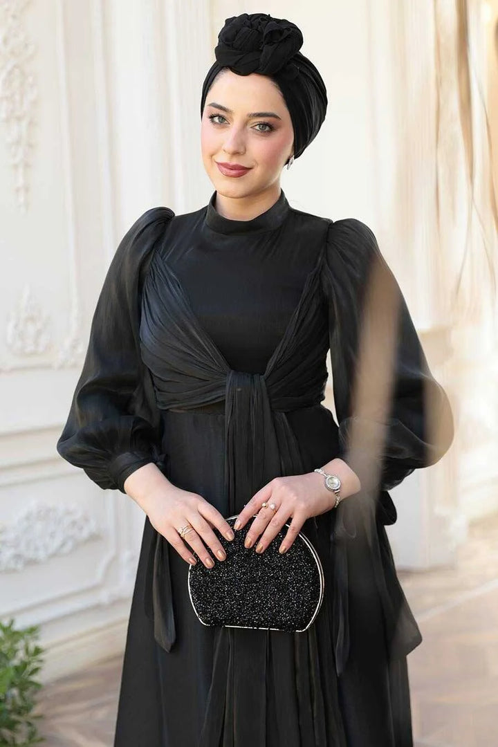 Woman wearing a Black Organza Criss Cross Gown with a glittery clutch, standing elegantly in a classic setting.