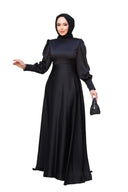 Woman wearing Black Satin Gown with fit and flare silhouette, crew neckline, and long sleeves, holding a small purse. Made in Turkey edition.