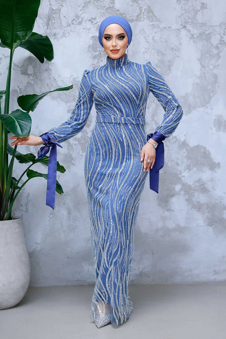 Woman wearing Blue Beaded Gown with crew neckline and long sleeves, standing against a gray textured wall with potted plant.