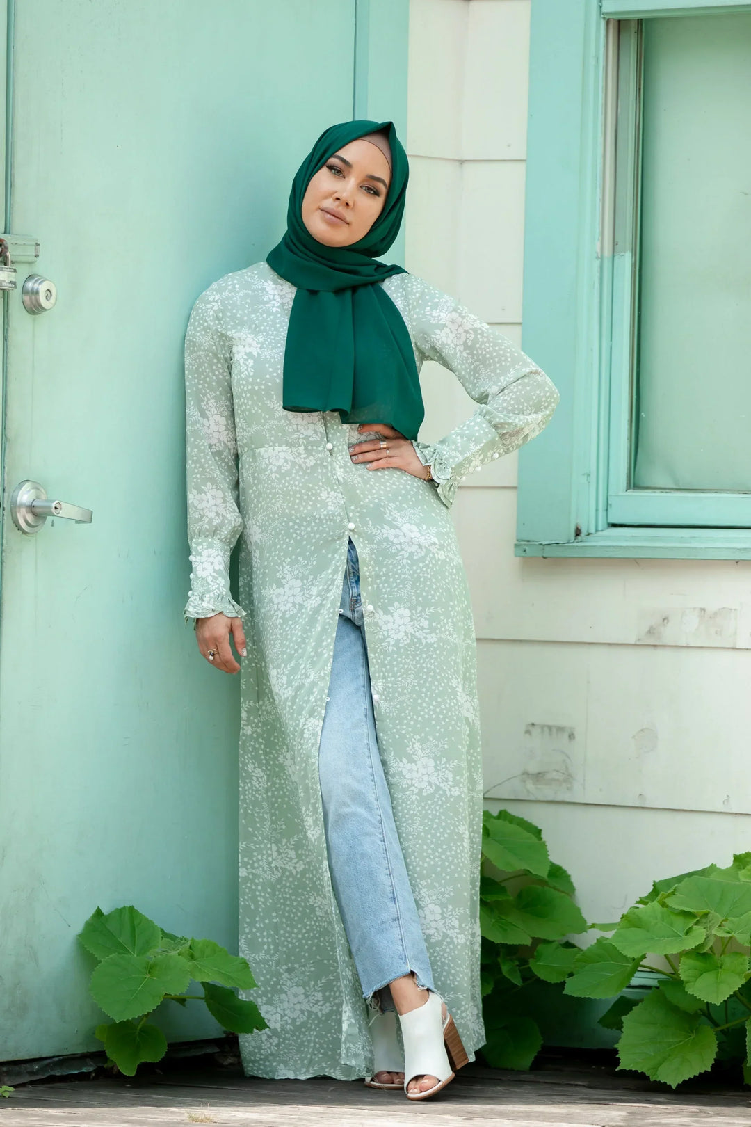 Woman wearing Forest Green Chiffon Hijab with a light green floral dress and jeans, standing by a turquoise door.