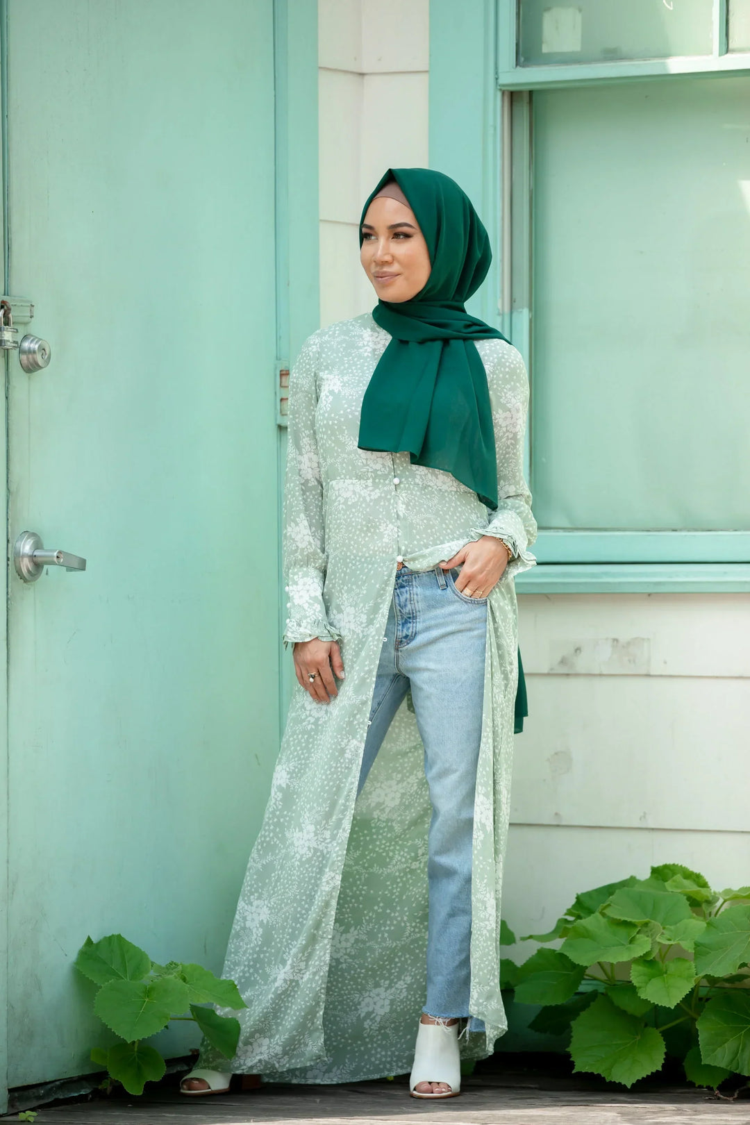 Woman wearing Forest Green Chiffon Hijab styled with a floral long shirt and jeans, standing by mint-colored door.