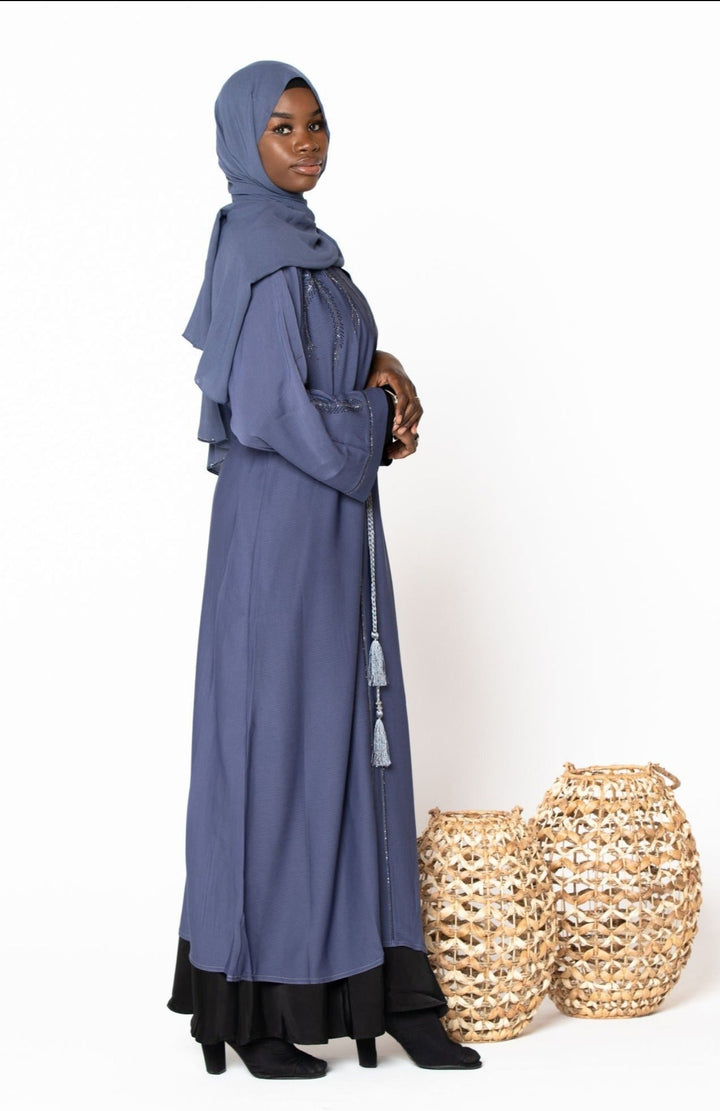 Woman in a gray Jamilah Sequin Open Front Abaya with drawstring tie, standing near woven baskets.