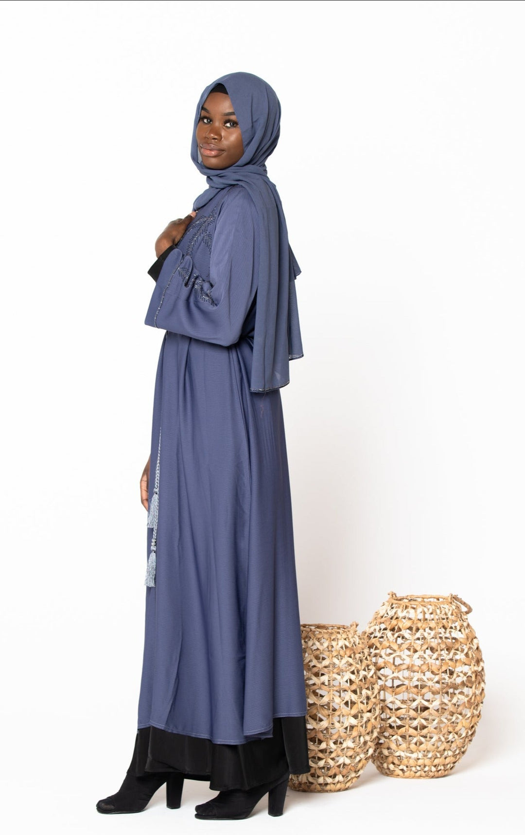 Woman wearing Gray Jamilah Sequin Open Front Abaya with hijab standing beside decorative baskets.