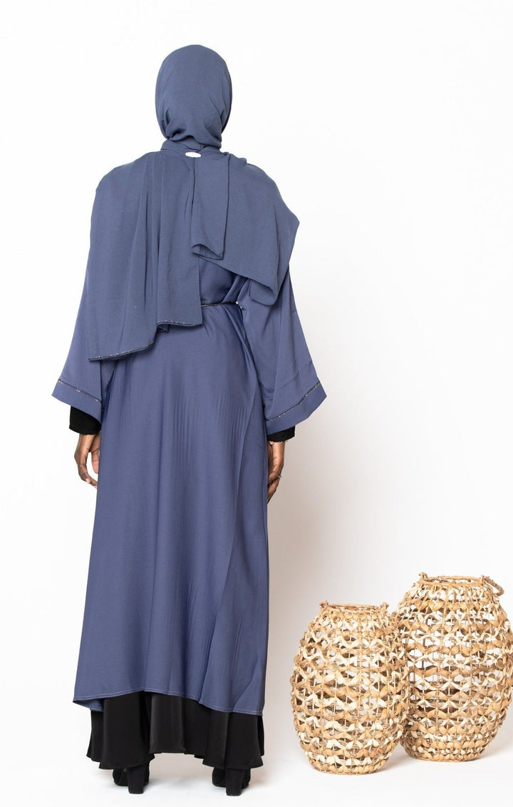 Back view of a woman wearing a gray Jamilah Sequin Open Front Abaya with a hijab, alongside two decorative baskets.