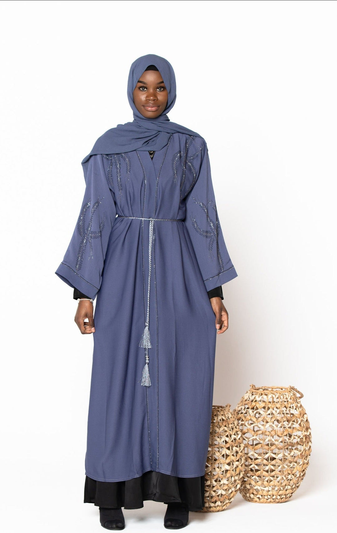 Woman wearing Gray Jamilah Sequin Open Front Abaya with sequin detailing and fabric belt, standing next to woven baskets.