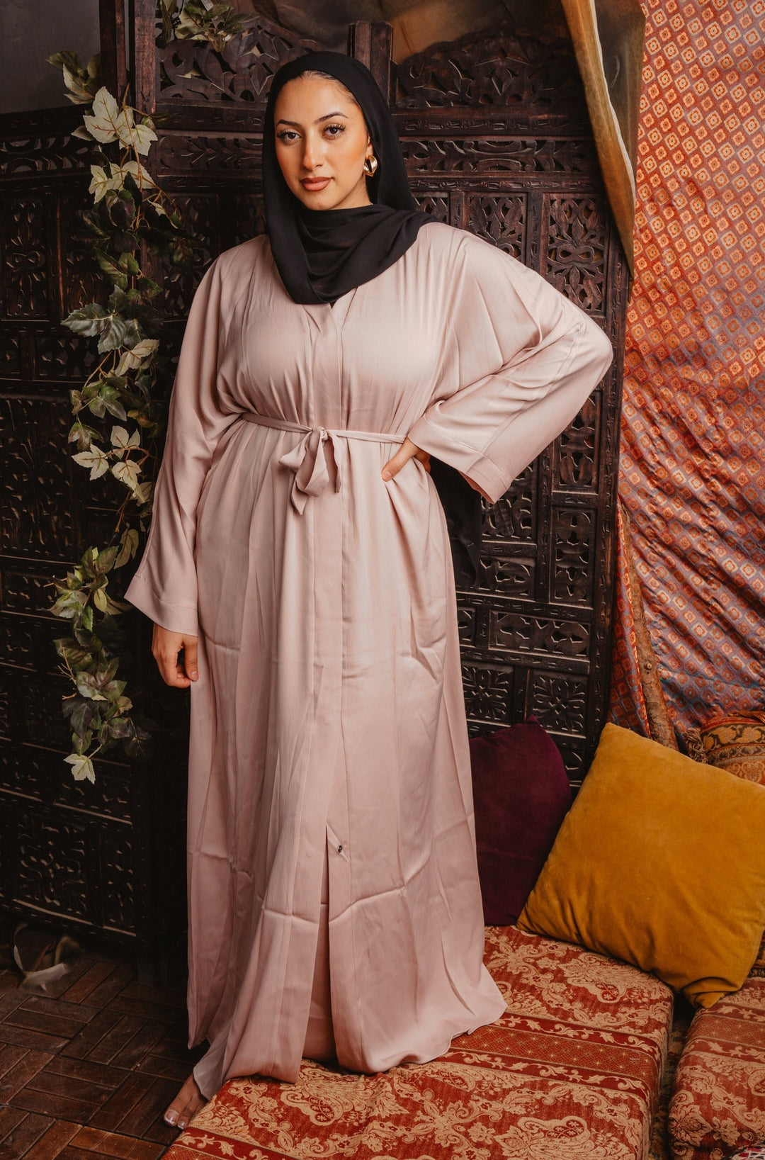 Woman wearing a Khaki Sleeveless Dress and Open Abaya Set with a black headscarf, standing in a room with ornate wood panelling and colorful cushions.