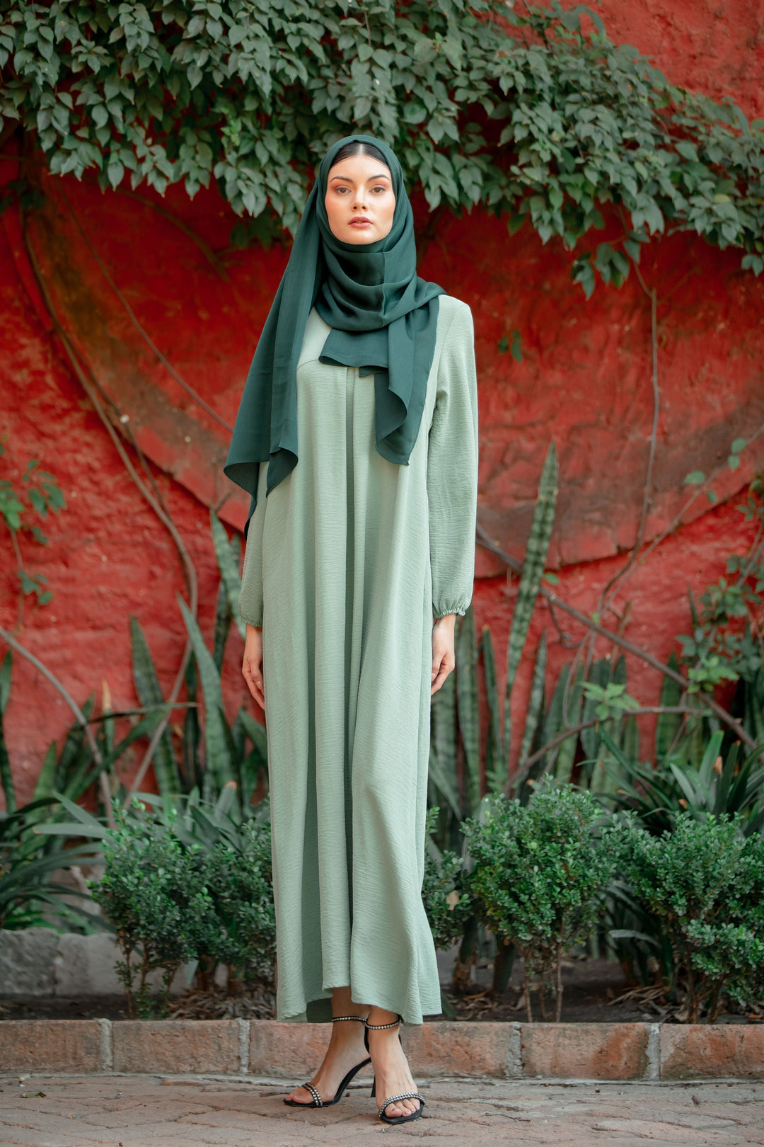 Woman wearing Mint Sheath Abaya Maxi Dress with kimono sleeves and fabric tie belt, posing outdoors against a lush, green, and red backdrop.