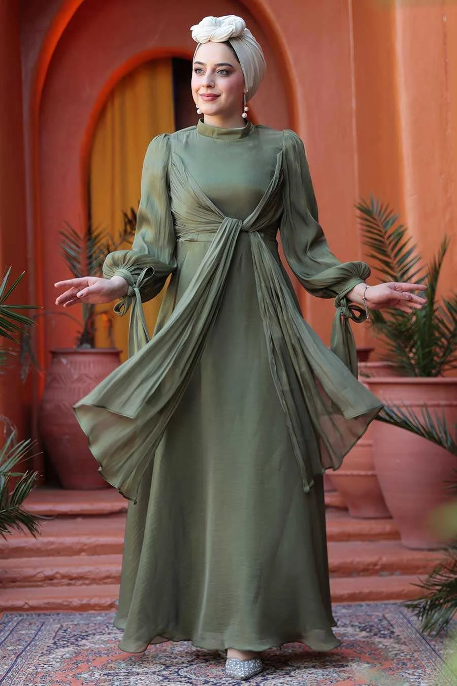 Woman wearing Olive Green Organza Criss Cross Gown with elegant fit and flare design, standing in front of a terracotta backdrop.
