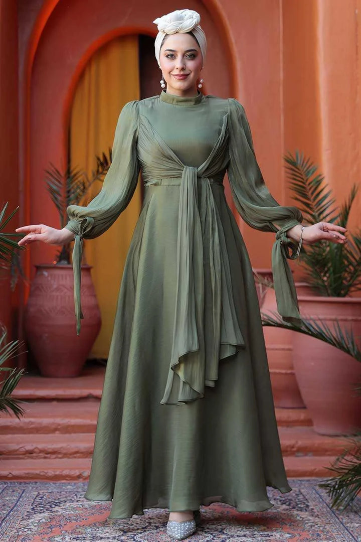 Woman wearing Olive Green Organza Criss Cross Gown with long sleeves and hijab, standing in front of terracotta building entrance.