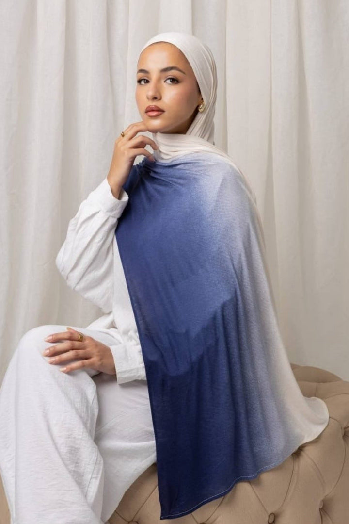 Woman wearing an Ombré Jersey Hijab in dark blue and white, sitting on a beige surface with a light curtain backdrop.