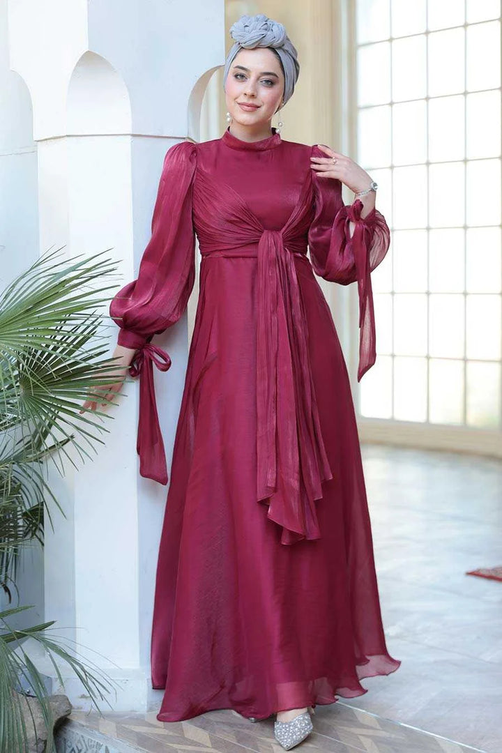 Woman wearing Red Organza Criss Cross Gown by Urban Modesty, featuring fit and flare silhouette with long sleeves, styled in a natural lit room.