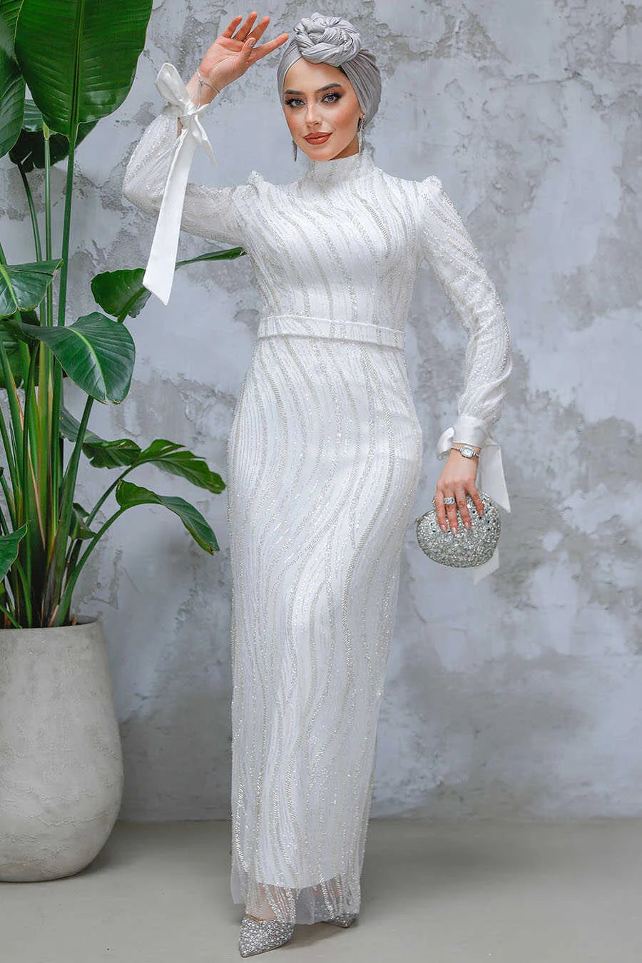 Woman wearing a White Beaded Gown with intricate detailing, standing against a textured wall, holding a sparkling clutch.