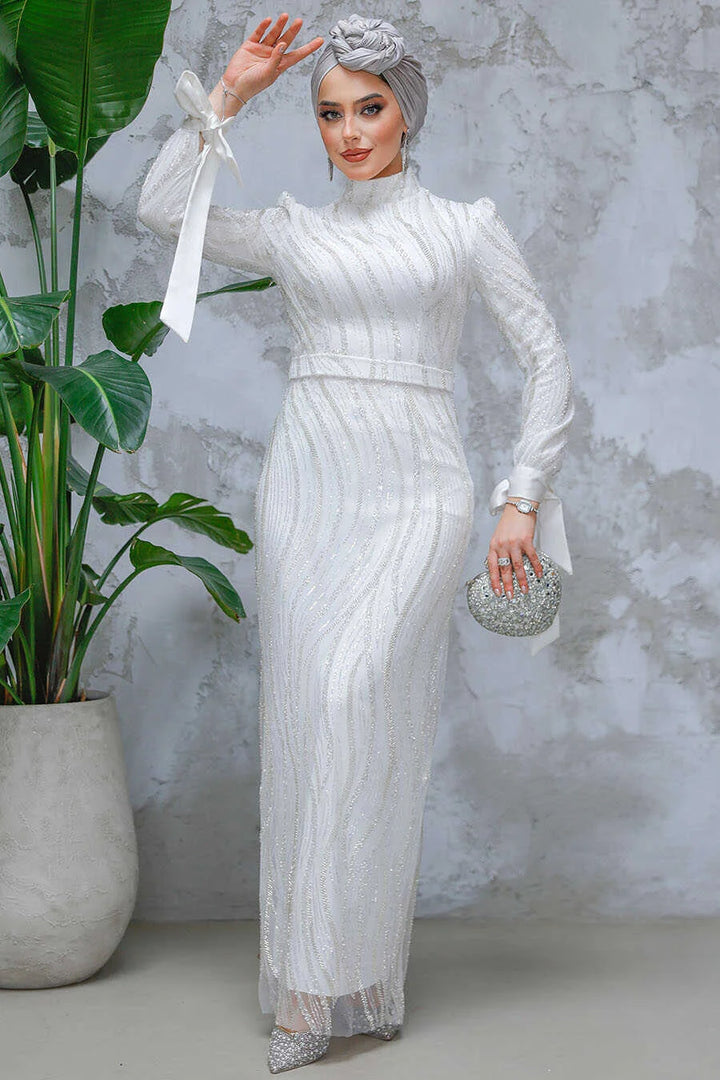 Woman wearing a White Beaded Gown with intricate detailing, standing against a textured wall, holding a sparkling clutch.