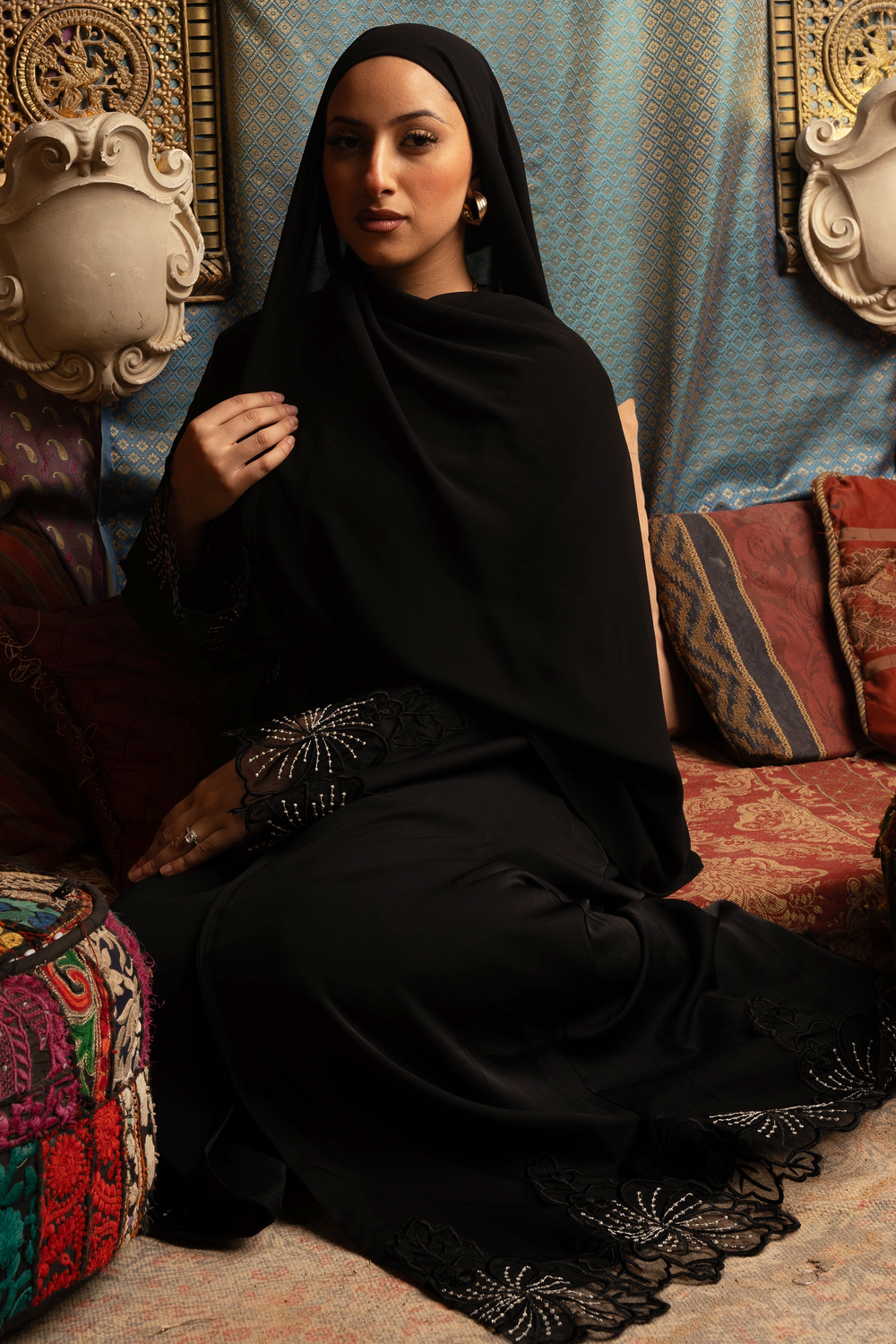 Elegant woman in a black lace sleeve abaya, adorned with kimono sleeves and sitting in a richly patterned setting.