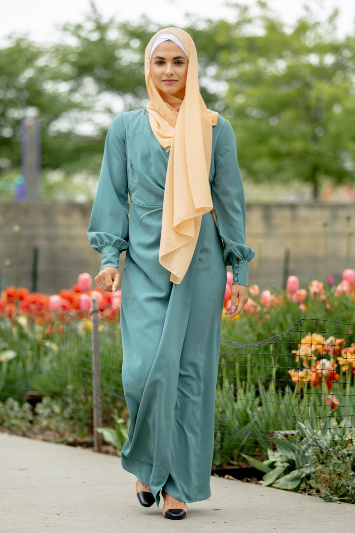 Woman in teal dress and peach hijab showcasing a sea blue wrap maxi dress with criss cross neckline